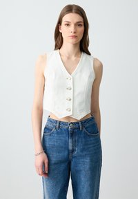 Top bianco corto con scollatura a V, caratterizzato da cinque bottoni di perla e orlo a punta. Abbinato a jeans in denim blu a vita alta.