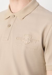Beige polo shirt lavet af tekstureret stof, med en to-knaps placket og et præget logo på brystet. Korte ærmer med syet kant.