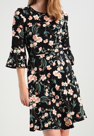 Frau trägt ein schwarzes Blumenkleid mit pinken Blumen, grünen Blättern, Spitzendetails an dreiviertel Ärmeln und einer taillierten Taille.