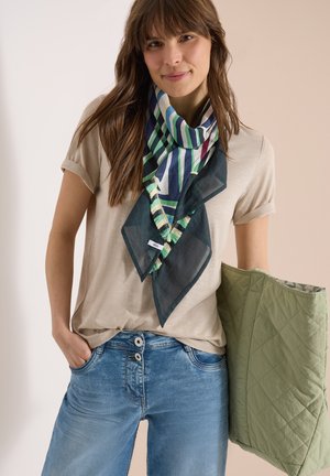 Nainen, joka pukeutunut beigeen t-paitaan ja sinisiin farkkuihin raidallisen huivin kanssa, pitää kädessään suurta tikattua vihreää olkalaukkua neutraalilla taustalla.