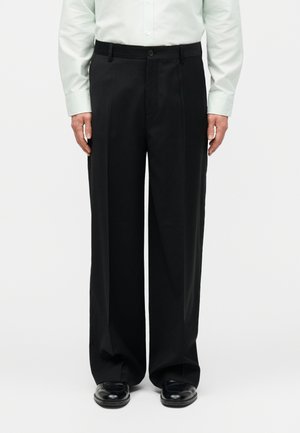 Άνδρας που φοράει μαύρο παντελόνι φαρδιάς γραμμής, λευκό πουκάμισο με κουμπιά και μαύρα εσώρουχα, στέκεται μπροστά σε ένα απλό λευκό φόντο.