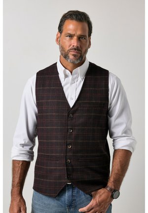 Mann trägt eine schwarz-rot karierte Weste über einem weißen Hemd mit hochgekrempelten Ärmeln und blauen Jeans, steht mit einer Hand in der Tasche.