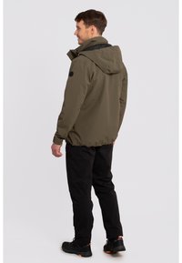 Olive green waterproof jacket with a black hood, adjustable hem, and side pockets, paired with black pants and dark shoes.