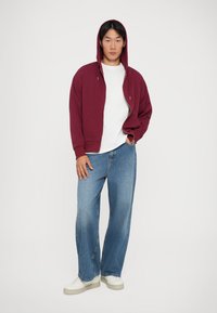 Jeune homme portant un hoodie zippé bordeaux, un t-shirt blanc, un jean bleu ample et des baskets blanches, se tenant avec une main dans sa poche.
