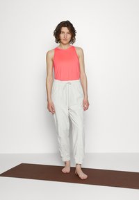 Top de alças cor-de-rosa com decote redondo, combinado com calças de jogging brancas elásticas com cordão na cintura. De pé sobre um tapete castanho.