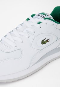 Sneaker bianca con accenti verdi, caratterizzata da una tomaia in pelle testurizzata, punta tonda, chiusura con lacci e un evidente patch con logo sulla linguetta.