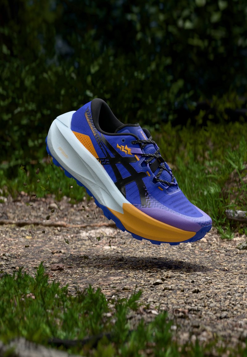 Chaussure de trail running avec un schéma de couleurs violet, noir et orange. Tige texturée, légère, avec un motif de semelle proéminent.