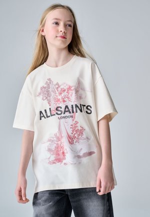Mladé dievča s dlhými blond vlasmi, ktoré nosí biele oversize tričko s ružovou potlačou anjela a textom "ALL SAINTS LONDON", v kombinácii s tmavými džínsami.