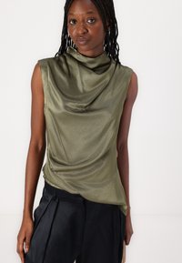 Haut sans manches vert olive avec un décolleté drapé et une texture lisse et brillante ; associé à un pantalon plissé sombre.