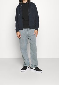 Sudadera con cremallera color navy con bolsillos delanteros sobre una camiseta negra, combinada con jeans rectos de lavado claro y zapatillas negras con detalles blancos.