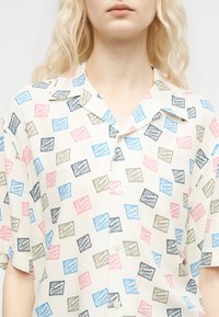 Femeie purtând o cămașă cu mânecă scurtă, cu nasturi, având un model geometric colorat repetitiv și un text care spune „DRAN Amour HOTEL.”