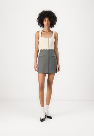 Robe bicolore avec décolleté carré ; partie supérieure crème et jupe grise avec deux poches avant et un design ajusté. Chaussures noires et chaussettes blanches.