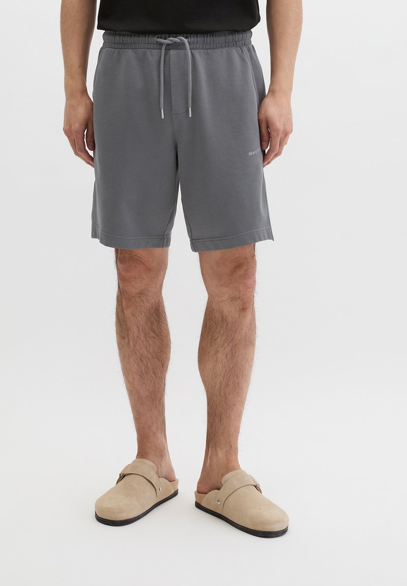 Mann trägt graue Shorts mit Kordelzug und beige Slip-On-Schuhe, steht vor weißem Hintergrund.