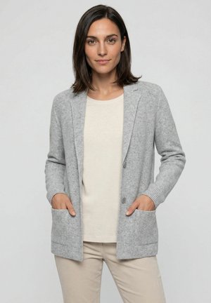 Vrouw staand met handen in de zakken, gekleed in een lichtgrijze cardigan, beige blouse en beige broek tegen een effen achtergrond.