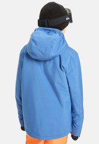 Personne portant une veste à capuche bleue, un bonnet en tricot noir, des gants noirs, un pantalon orange et des lunettes de ski noires, de dos par rapport à l'appareil photo.