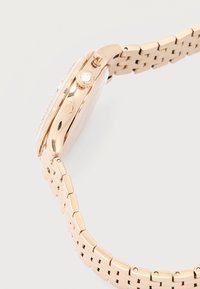 Ροζ χρυσό ρολόι χειρός, πλευρική όψη που δείχνει λεπτομερώς το κουμπί ρύθμισης με ενσωματωμένη διαφανή πέτρα και μεταλλικό μπρασελέ με συνδέσμους σε λευκό φόντο.