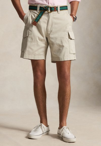 Homme portant un short cargo beige avec des poches à rabat, une ceinture en tissu rayée, des baskets blanches et une chemise rose retroussée dans un cadre neutre.