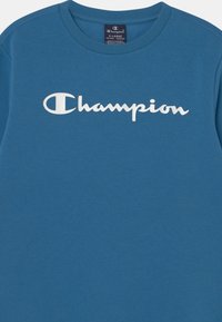 Modré bavlněné tričko s kulatým výstřihem, na prsou velké bílé logo "Champion" a strukturovaný povrch látky.