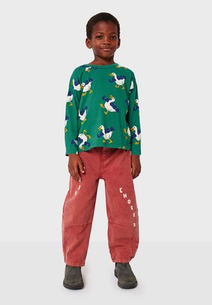 Chemise verte à manches longues avec des canards blancs et du texte bleu, associée à un pantalon en velours côtelé rouge avec des lettres blanches sur le côté. Chaussures grises.