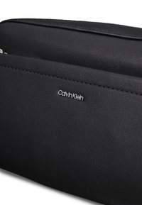 Sort syntetisk håndtaske med en glat tekstur, der har et sølvfarvet Calvin Klein-logo, et slankt design og præcise syningsdetaljer.