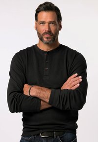 Schwarzes Langarm-Henley-Shirt aus strukturiertem Stoff, mit Rundhalsausschnitt und einer Dreiknopf-Leiste, getragen mit dunklen Jeans.