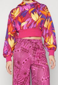 Lila huvtröja med blommönster, har långa puffärmar och ett rosa band; kombinerad med rosa mönstrade byxor, elastisk midja.