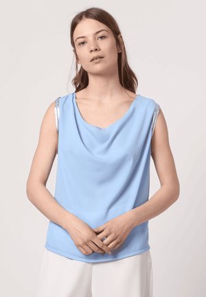 Mujer de pie, con un top azul claro sin mangas y escote drapeado, y pantalones blancos; manos entrelazadas, fondo neutro.