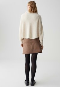 Cremefarbener gerippter Strickpullover, brauer Wildleder-Minirock, schwarze Strumpfhosen und schwarze Schuhe; Fokus auf Textur und geschichtetes Design.