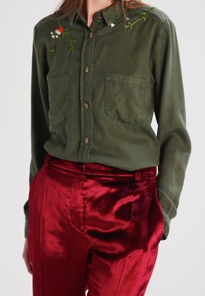 Camicia verde oliva con ricami floreali, chiusura frontale con bottoni e due tasche sul petto, abbinata a pantaloni di velluto rosso lucido.