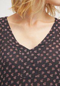 Tissu noir avec de petits motifs floraux rouges, doté d'un décolleté en V et de plis subtils à la poitrine, créant un aspect texturé.