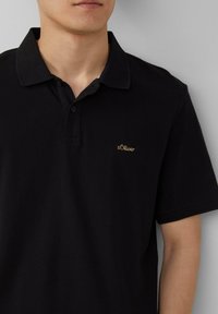 Schwarzes Polohemd aus strukturiertem Stoff, mit einem kragenförmigen Ausschnitt, zwei Knöpfen und einem kleinen goldenen gestickten Logo auf der linken Brust.