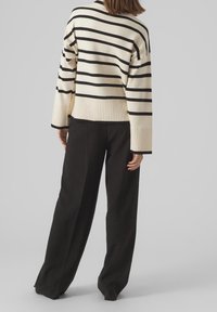 Randig tröja i krämfärgad och svart med en avslappnad passform, ribbad nederkant och vida ärmar, tillsammans med svarta vida byxor mot en grå bakgrund.