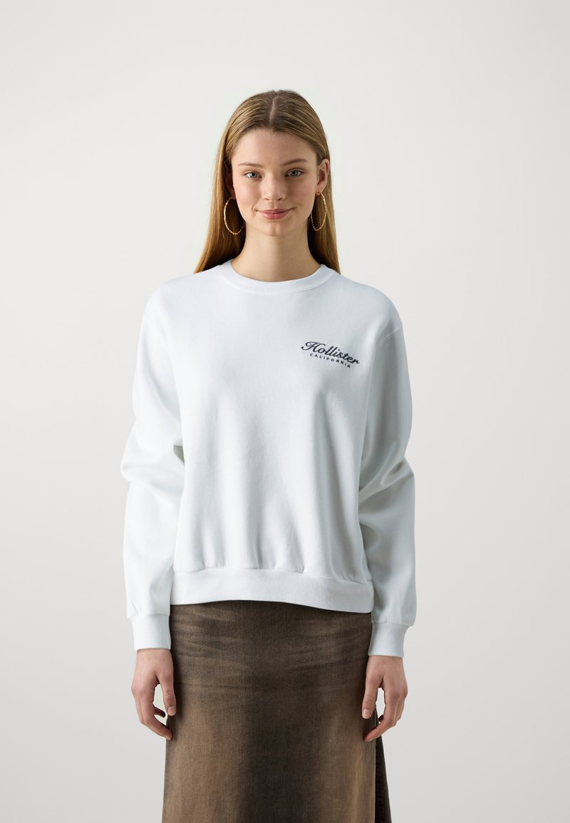 Λευκή μπλούζα με στρογγυλή λαιμόκοψη, ριμπ μανσέτες και στρίφωμα; διαθέτει μαύρο λογότυπο στο στήθος. Το μοντέλο φοράει μια καφέ φούστα φτιαγμένη από υφασμάτινη υφή.