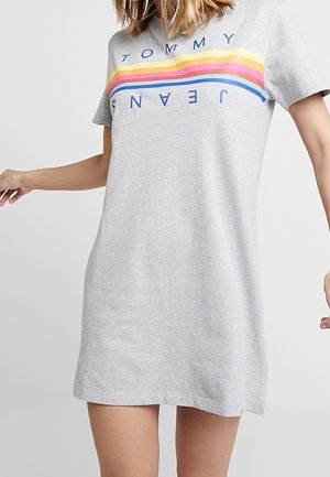 Kobieta w jasnoszarej sukience z krótkim rękawem z poziomymi tęczowymi paskami i napisem „TOMMY JEANS” na piersi.