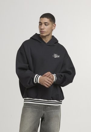 Czarna bluza z kapturem z białym haftowanym logo, ściągaczami na rękawach i u dołu z białymi paskami, noszona z szarymi dżinsami.