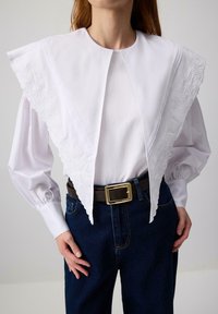 Donna che indossa una camicia bianca con un ampio colletto ricamato e maniche lunghe, abbinata a jeans blu scuro e una cintura nera con fibbia dorata.