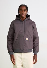 Gray zip-up hoodie with a lined hood, front pocket, and ribbed cuffs. Features a small orange Carhartt logo patch on the left pocket.
