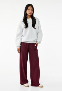 Grå oversize sweatshirt med V-ringning, parat med vida vinröda byxor. Modellen står framför en helt vit bakgrund, iförd casual sneakers.