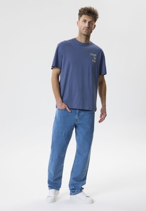 Egy férfi áll, kék pólót visel „c’est la vie” felirattal, világoskék farmert és fehér tornacipőt egy egyszerű világos háttér előtt.