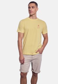 Žluté bavlněné tričko s kr átkým rukávem a kulatým výstřihem, s malou grafikou slunce a vlny v červené barvě na levé straně hrudníku.