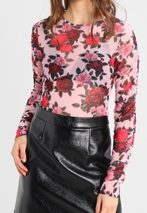 Kobieta ubrana w przezroczystą różową bluzkę z długim rękawem z nadrukiem czerwonych i różowych róż oraz czarną błyszczącą skórzaną spódnicę.