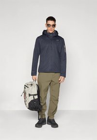 Marinblå huvjacka i lättviktigt material med dragkedja fram. Olivgröna byxor med cargofickor, kombinerade med en grå ryggsäck och svart skor.