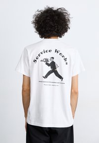 White organic cotton t-shirt featuring a black graphic of a waiter on the back, with "Service Works" text above and below the illustration.