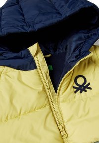 Prošívaná bunda se žlutým tělem a námořnicky modrou kapucí, s zipovým zapínáním a vyšitým logem na hrudi. Hladký vnější povrch.