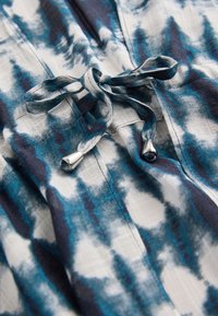 Niebiesko-biała tkanina z wzorem, ozdobiona sznurkiem z srebrnymi akcentami. Materiał wydaje się lekki o fakturowanej powierzchni.