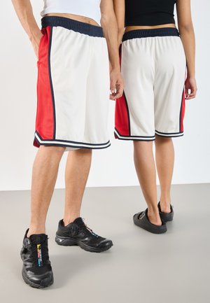 Bermudas deportivas blancas y rojas con acentos en azul marino; cuentan con bolsillos laterales, cintura elástica y una textura de malla. Llevadas con zapatillas negras.