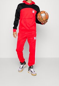 Sweat à capuche rouge avec des accents noirs et des rayures blanches, assorti à un pantalon de jogging rouge. Tient un ballon de basket, porte des baskets noires et blanches.