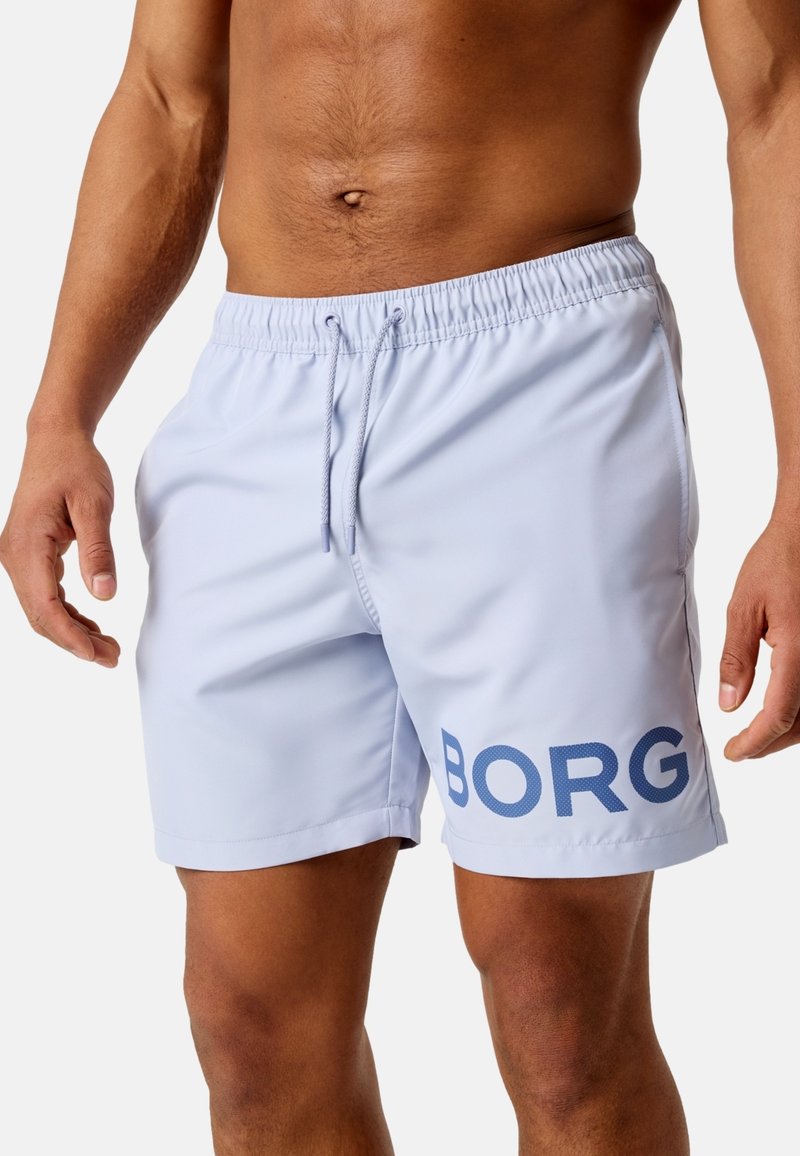 Männlicher Torso, der hellblaue Badeshorts mit dem dunkelblau aufgedruckten Schriftzug „BORG“ am linken Bein vor einem einfarbigen Hintergrund trägt.
