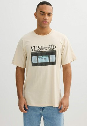 Joven con camiseta beige con un gráfico de cinta VHS etiquetado como "My Hard Life 1998" y texto "VHS Very Human Stories" posando frente a un fondo blanco.
