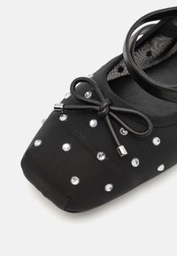 Black ballet flat made of textured fabric, adorned with reflective rhinestones, featuring a bow detail and a thin strap.